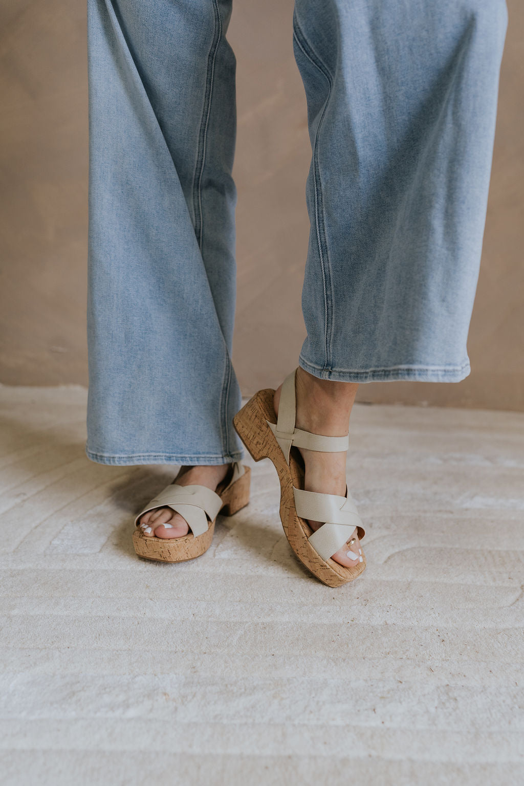 Front view of female model wearing the Dasha Sandal in Off White which features off white upper leather fabric, kork platform sole, block heel, criss-cross strap and gold adjustable buckle closure