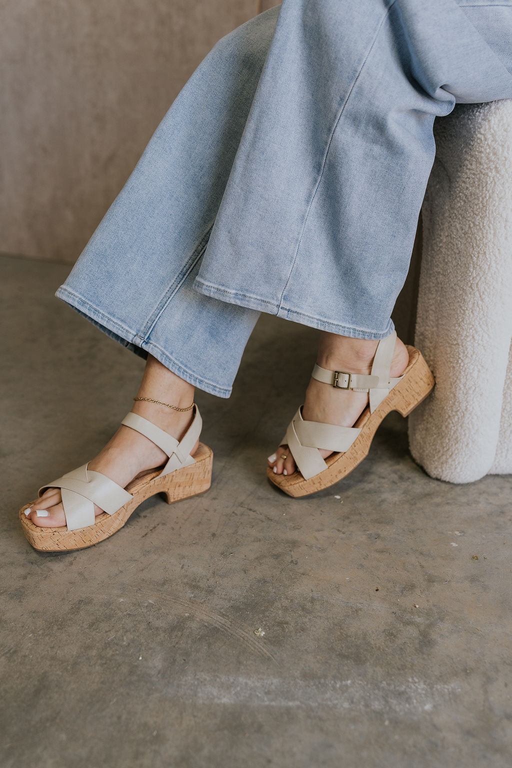 Left side view of female model wearing the Dasha Sandal in Off White which features off white upper leather fabric, kork platform sole, block heel, criss-cross strap and gold adjustable buckle closure