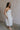 Full body side view of female model wearing the Rebecca Off White Pearl Bow Midi Dress which featuresCream Sheer Fabric, Midi Length, Cream Lining, Strapless, Pearl Details and Bow Back Detail