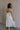 Full body back view of female model wearing the Rebecca Off White Pearl Bow Midi Dress which featuresCream Sheer Fabric, Midi Length, Cream Lining, Strapless, Pearl Details and Bow Back Detail