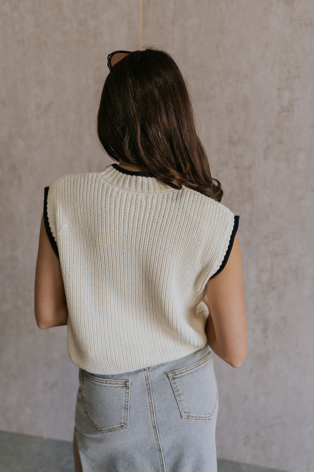 Back view of female model wearing the Vivian Ivory & Navy Knit Sleeveless Sweater which features Cream Cable Knit Fabric, Navy Trim Details, Left Front Chest Pocket, High Neckline and Sleeveless.