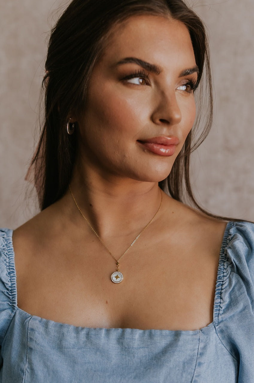 Close up view of female model wearing the Angelica White & Gold Star Medallion Necklace which features adjustable gold chain layer with an ivory circle medallion, gold star design and rhinestones around the medallion