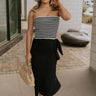 Full body front view of model wearing the Arabella Black Wrap Midi Skirt that has black lightweight fabric, a wrap front with a side tie detail, and back slit. Worn with striped tube top.