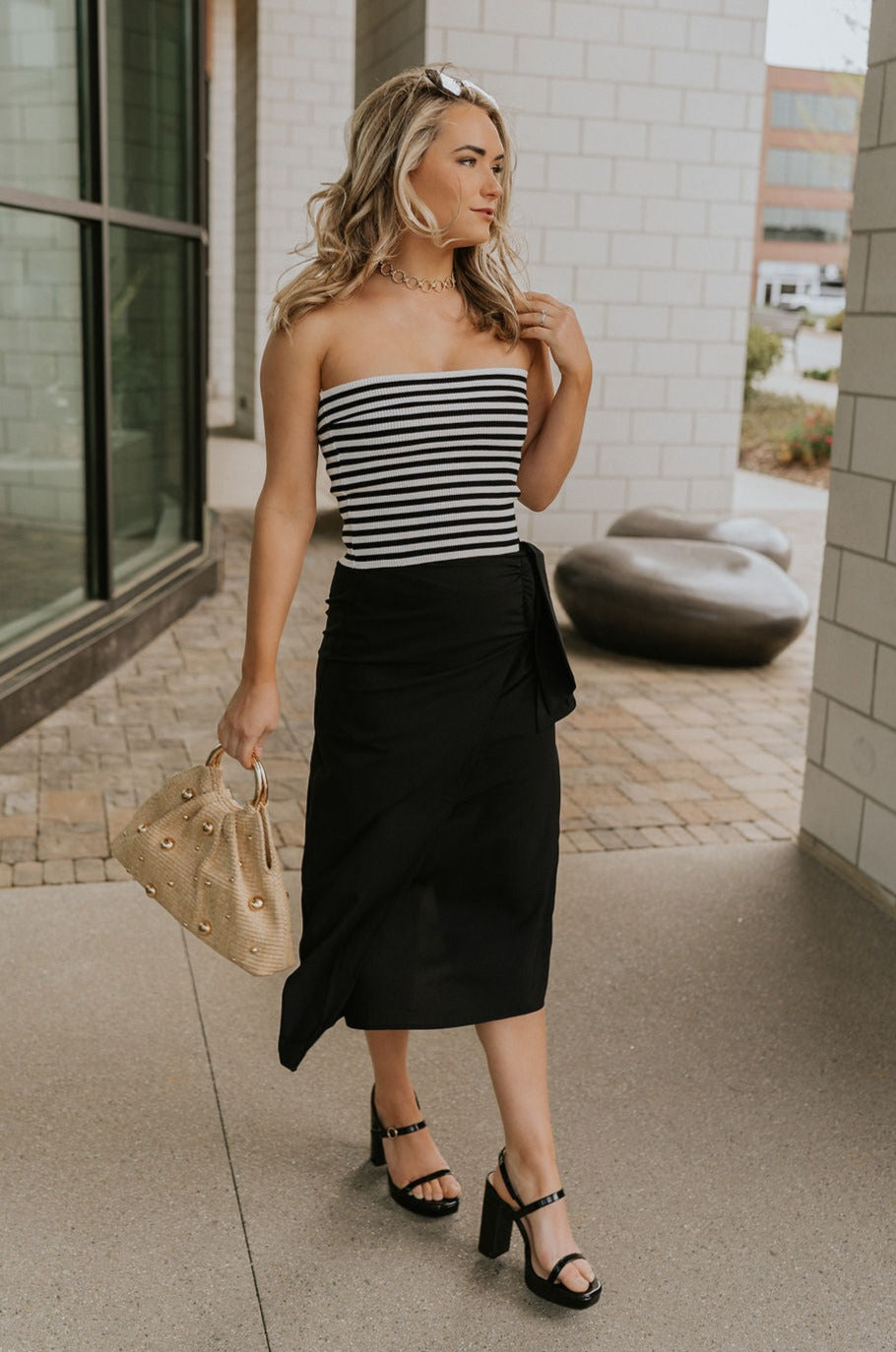 Full body front view of model wearing the Arabella Black Wrap Midi Skirt that has black lightweight fabric, a wrap front with a side tie detail, and back slit. Worn with striped tube top.