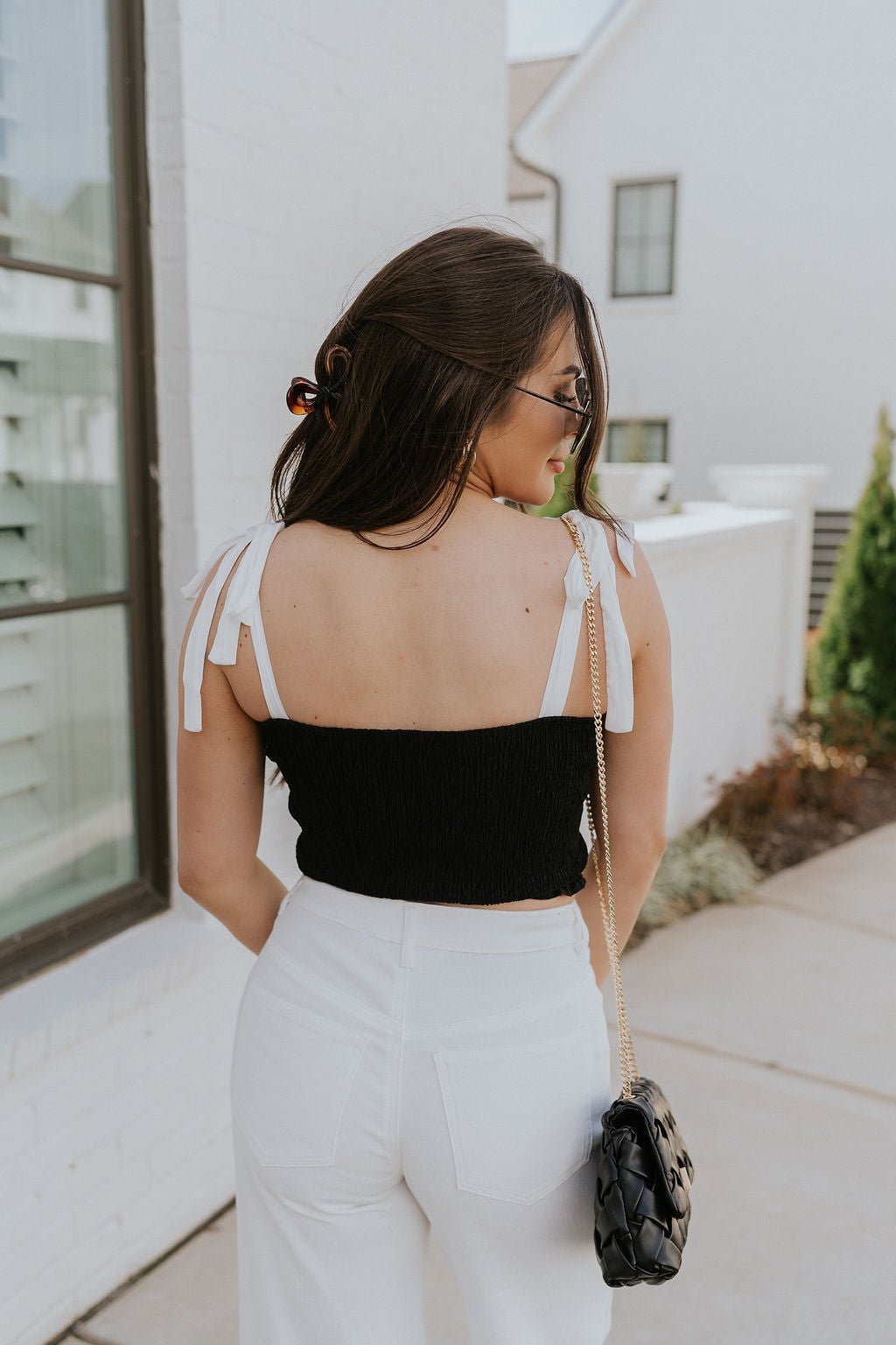 Back view of female model wearing the Reese Black & White Cropped Tank Top that has black smocked body, white tie straps and a cropped waist. Worn with white pants.