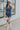 Full body front view of female model wearing the Georgia Belted Denim Romper in medium wash, that has a button up front, collar, and belted waist. Paired with white boots.