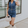 Full body front view of female model wearing the Georgia Belted Denim Romper in medium wash, that has a button up front, collar, and belted waist. Paired with white boots.