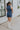 Full body back view of female model wearing the Georgia Belted Denim Romper in medium wash, that has a button up front, collar, and belted waist. Paired with white boots.