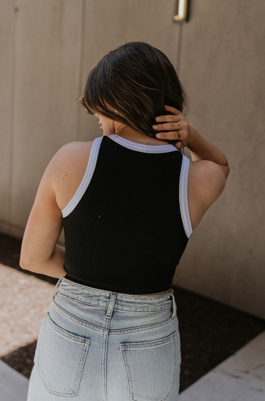 Upper body back view of female model wearing the Ari Black & White Brami that has black ribbed fabric with white trim and a round neck. Paired with blue jeans.