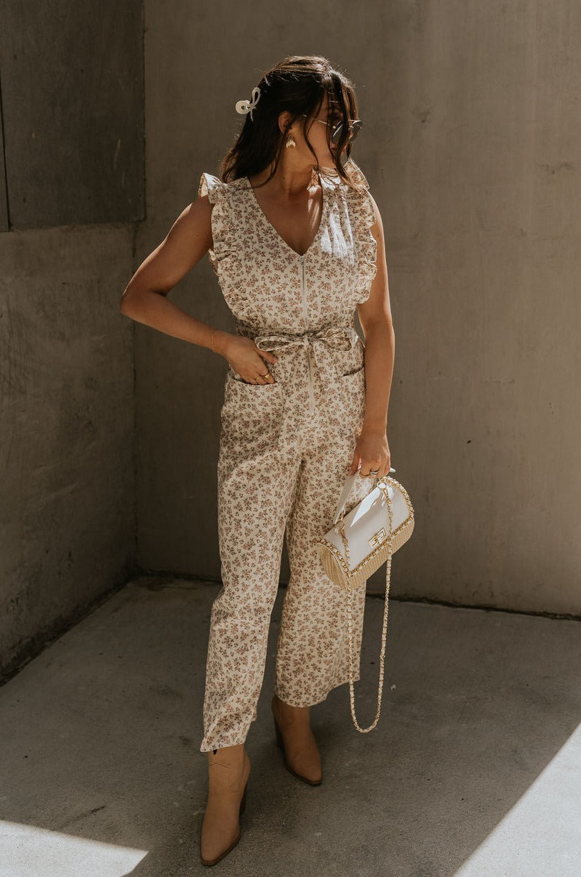 Full body view of female model wearing the Myla Cream Pink Floral Sleeveless Jumpsuit which features Cream Cotton Fabric, Pink Floral Print, Wide Leg Pants,  Tie Strap with Belt Loops, V-Neckline and Ruffle Straps