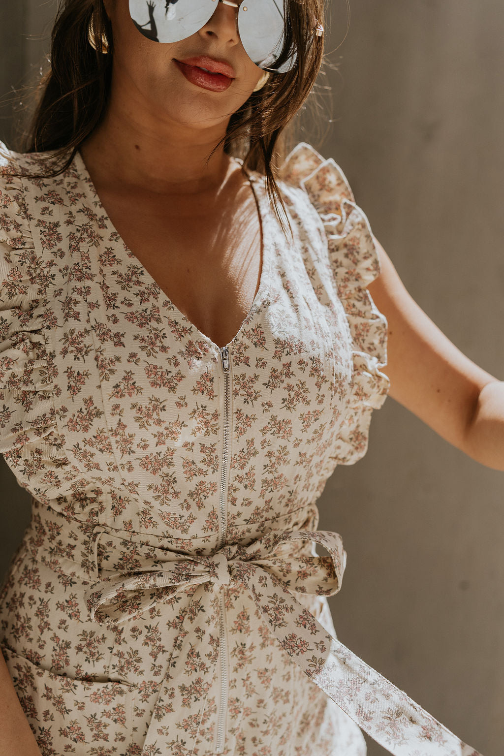 Close up view of female model wearing the Myla Cream Pink Floral Sleeveless Jumpsuit which features Cream Cotton Fabric, Pink Floral Print, Wide Leg Pants, Tie Strap with Belt Loops, V-Neckline and Ruffle Straps