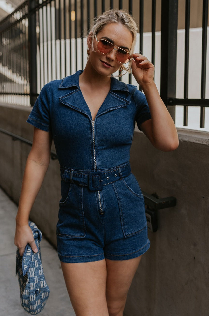 Front view of female model wearing the Mavis Denim Short Sleeve Zip-Up Romper which features Denim Fabric, Two Front Pockets, Front Zipper Closure, Adjustable Monochrome Belt with Loops, Collared Neckline and Short Sleeves