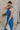 Side view of female model wearing the Juliette Blue Halter Tie Pleated Jumpsuit which features Blue Lightweight Fabric, Pleated Wide Pant Legs, Front Key Hole Design, Halter Neckline Tie Closure and Open Back