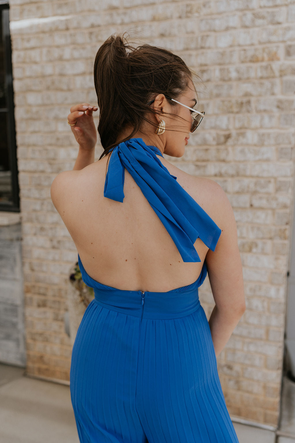 Back view of female model wearing the Juliette Blue Halter Tie Pleated Jumpsuit which features Blue Lightweight Fabric, Pleated Wide Pant Legs, Front Key Hole Design, Halter Neckline Tie Closure and Open Back