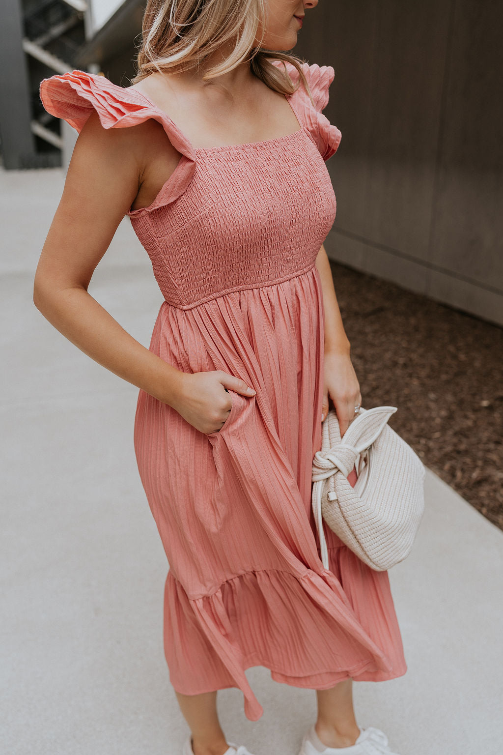 Side view of female model wearing the Allie Coral Pink Ruffle Midi Dress which features Coral Pink Lightweight Fabric, Midi Length, Coral Pink Lining, Ruffle Tier Skirt, Smocked Upper, Square Neckline, Ruffle Straps and Two Side Pockets