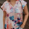 Close up view of female model wearing the Vanessa Watercolor Multi Short Sleeve Top which features Satin Fabric, Purple, Blue, Peach and Pink Watercolor Block Print, V-Neckline Cut-Out and Ruffle Short Sleeves