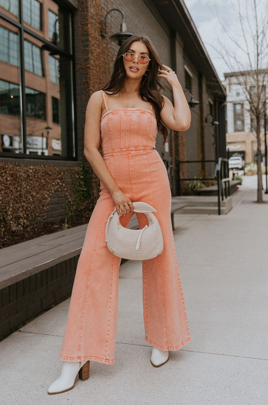 Full body front view of brunette model wearing the Regan Wide Leg Denim Jumpsuit in Coral, that has coral denim fabric, wide legs, and thin straps. Model is holding beige purse in front of her.
