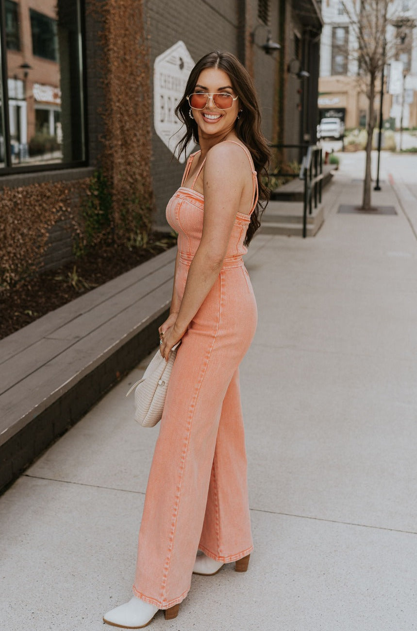 Full body side view of brunette model wearing the Regan Wide Leg Denim Jumpsuit in Coral, that has coral denim fabric, wide legs, and thin straps. Model is holding beige purse in front of her.
