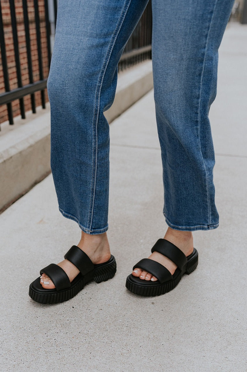 Side view of female model wearing the ONA Streetworks Slide Flat Sandal in Black & Chalk which features Black Light Foam Leather Upper, Two Straps, Slide-On Style, High-Traction Rubber Platform Sole and 2" Heel Height, 1 1/2" Platform Height