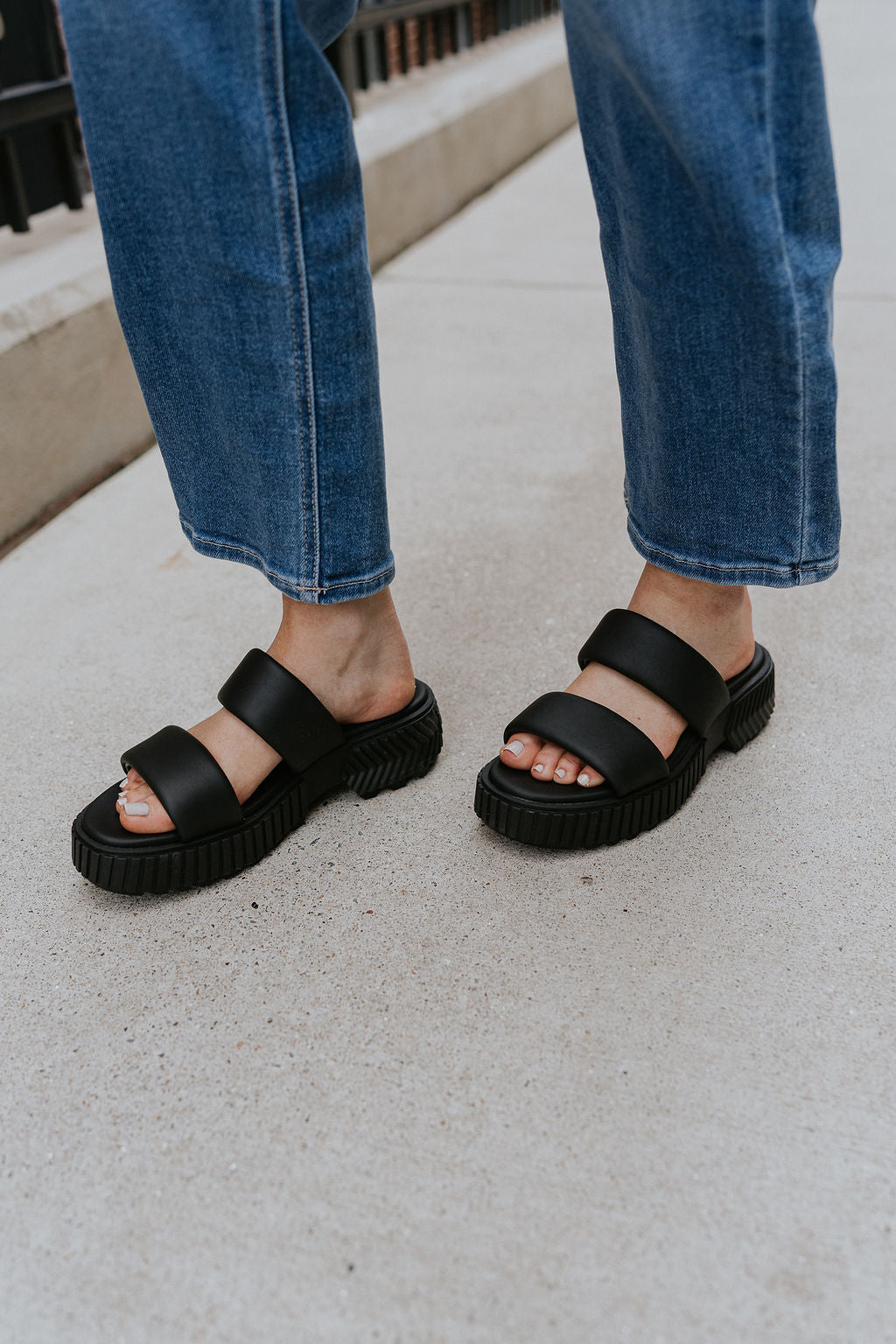 Side view of female model wearing the ONA Streetworks Slide Flat Sandal in Black & Chalk which features Black Light Foam Leather Upper, Two Straps,  Slide-On Style, High-Traction Rubber Platform Sole and 2" Heel Height, 1 1/2" Platform Height 