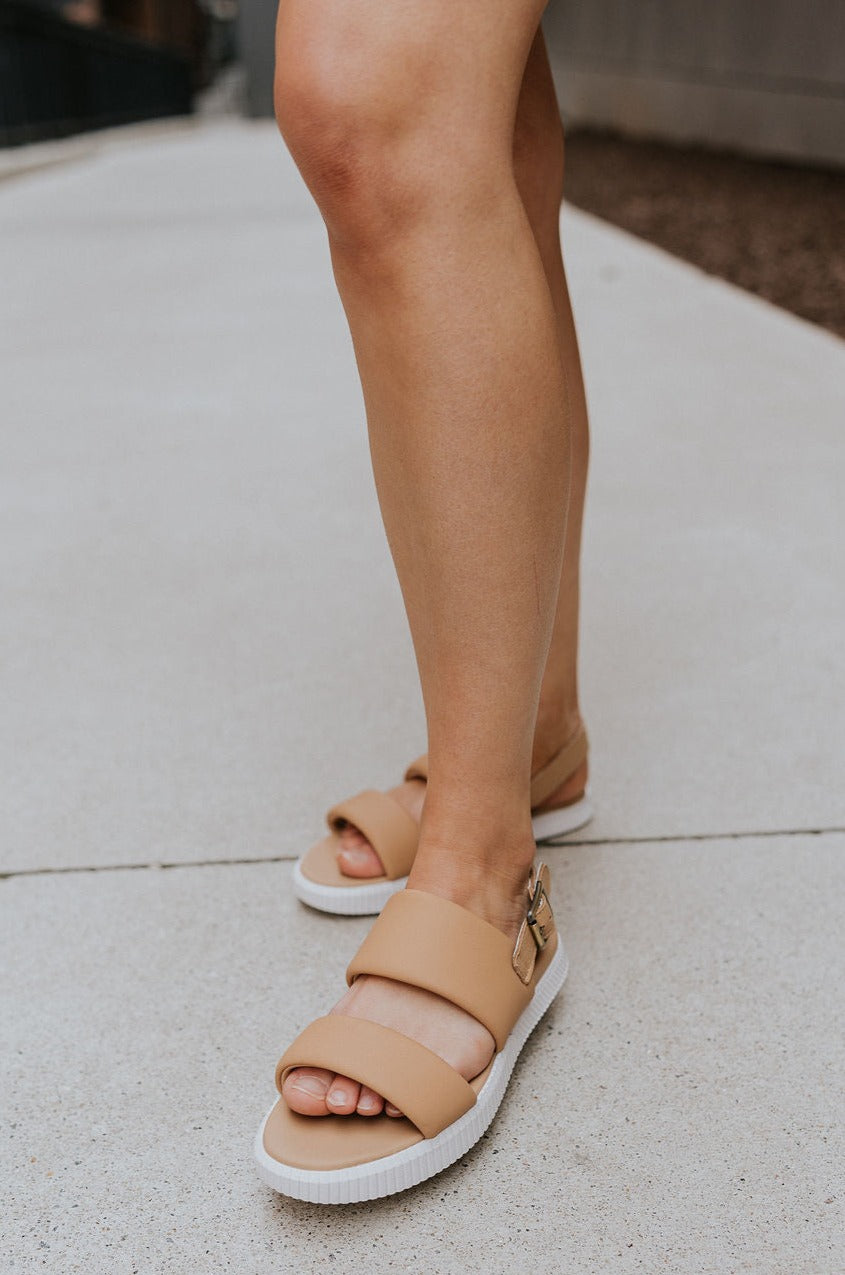 Frontal side view of female model wearing the ONA Streetworks Go-To Flat Sandal in Honest Beige & Sea Salt which features Beige Leather Upper, Adjustable Buckle Closure with Back Strap, White Molded Rubber Sole and 1" Platform Height