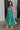Full body side view of female model wearing the Vera Halter Cross Neckline Midi Dress in Green which features Lightweight Fabric, Lining, Midi Length, Side Ties Waistband, Halter Cross Neckline, Adjustable Straps and Sleeveless.