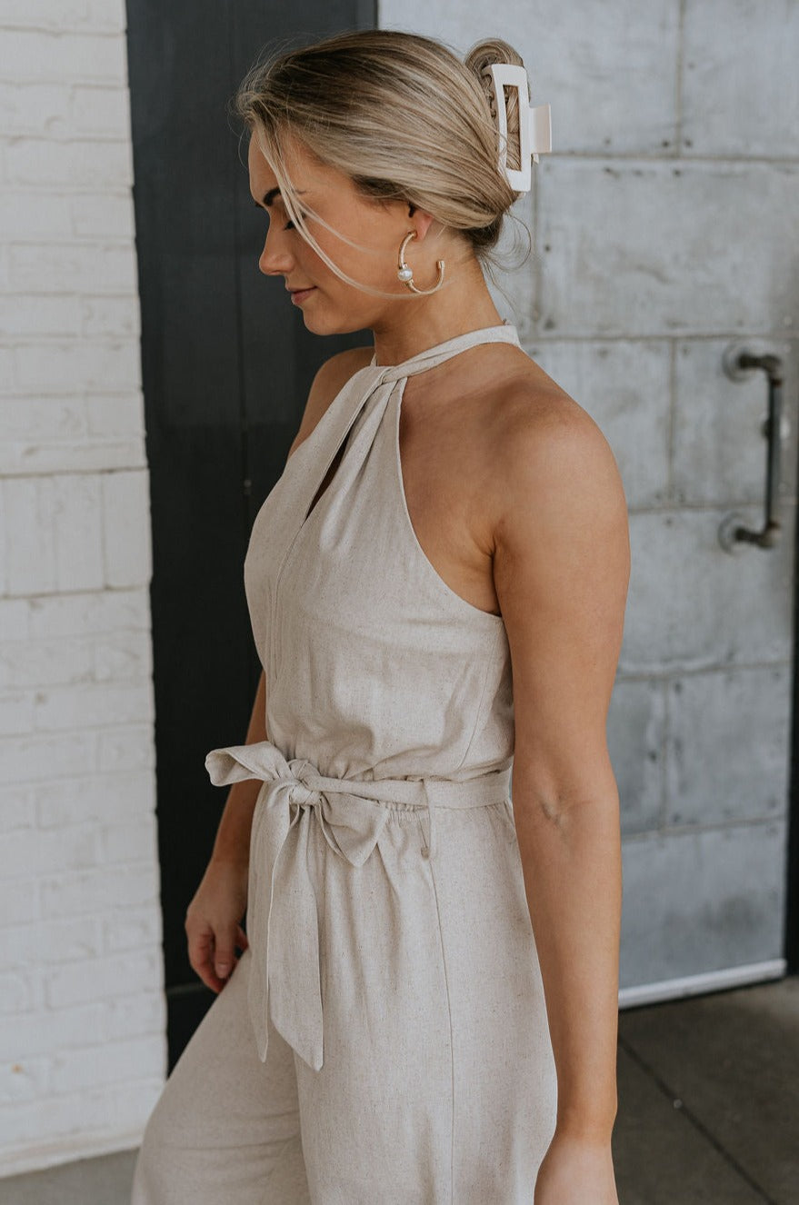 Side view of female model wearing the Lilah Natural Linen Halter Neckline Jumpsuit which features Natural Linen Fabric, Wide Pant Legs, Tie Waistband, Twist Halter Neckline with Button Closure and Back Key Hole Design