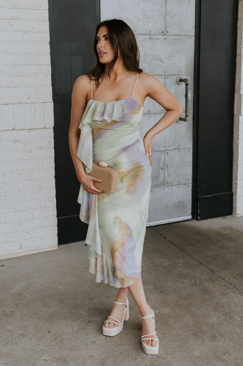 Full body view of female model wearing the Noelle Green Watercolor Multi Ruffle Midi Dress which features Mesh fabric with a green, purple, pink, and yellow watercolor pattern, Green lining, Midi Length, Ruffles across the neckline that drape down the right side, Left side ruching and Adjustable thin straps