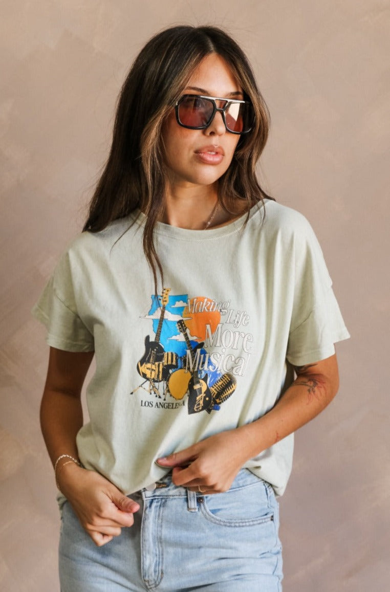 Front view of female model wearing the Los Angeles Musical Light Sage Graphic Top which features Light Sage Cotton Fabric, Short Sleeve Top, Round Neckline and Los Angeles, CA Musical Graphic