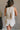 Back view of female model wearing the Elodie Beige Ruffle Tier Sleeveless Romper which features Beige Lightweight Fabric, Beige Lining, Ruffle Tier Details, Round Neckline, Sleeveless and Back Button Closure