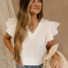 Front view of female model wearing the Jemma White Ruffle Top that has white fabric, short ruffled cap sleeves, a v neckline, and keyhole back. Worn with blue jeans.