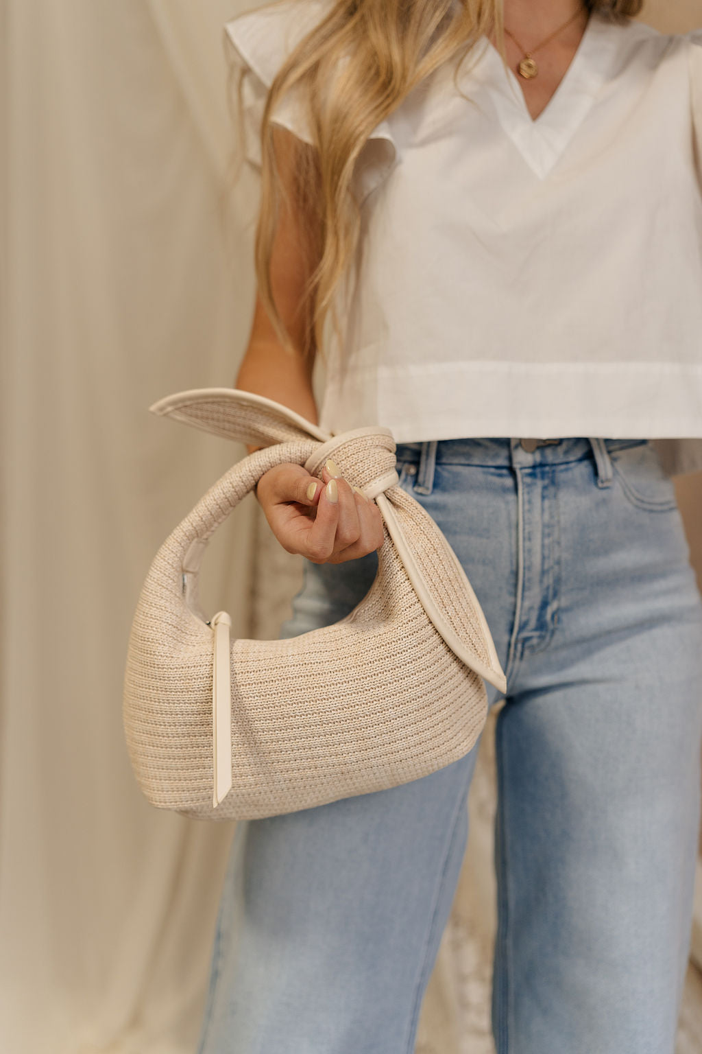 Close front view of model holding the Macy Ivory Knot Handbag. This features a ivory woven fabric with a knot handle. There is a zipper closure along with a pocket on the inside.