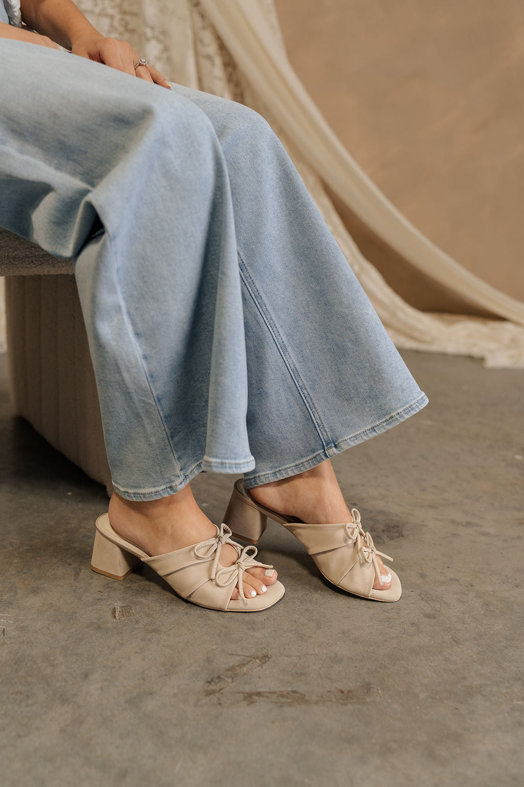 Side view of female model sitting; her legs are shown in jeans and the Emerald Light Grey Bow Ties Heel Sandal which features grey suede material, 2.5" block heels, backless slide entry, and ruched uppers with a cutout and bow details.
