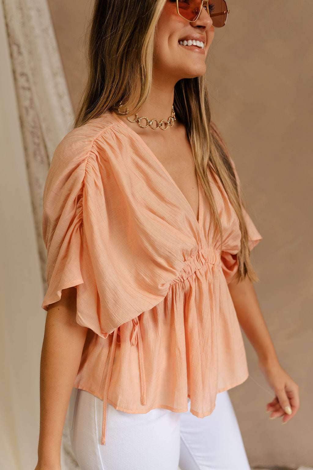 Side view of female model wearing the Juliet Peach Ruched Short Sleeve Top that features flowy peach fabric, ruched short sleeves, and an elastic waist.