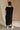 Full body back view of female model wearing the Remy Black Short Sleeve Midi Dress that features black knit fabric, short sleeves, midi length, and side slits. =