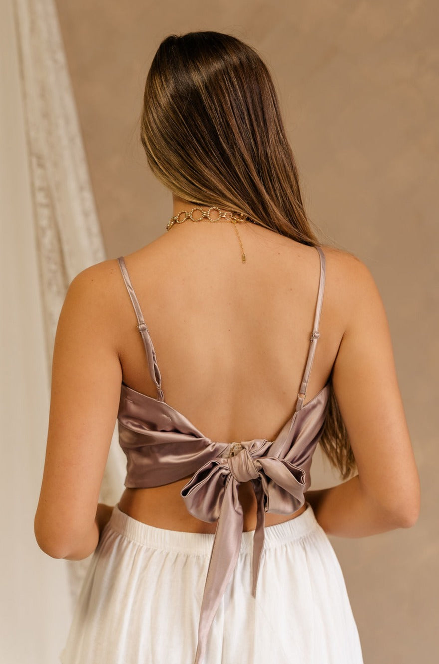 Back view of female model wearing the Jovie Mauve Satin Tank that has mauve satin fabric, thin straps, and a tie back. Paired with white skirt.