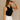 Side view of model wearing the Rayna Black Ribbed Tank, that features wide ribbed fabric, a round neckline, and sleeveless body. Worn with white shotrs.