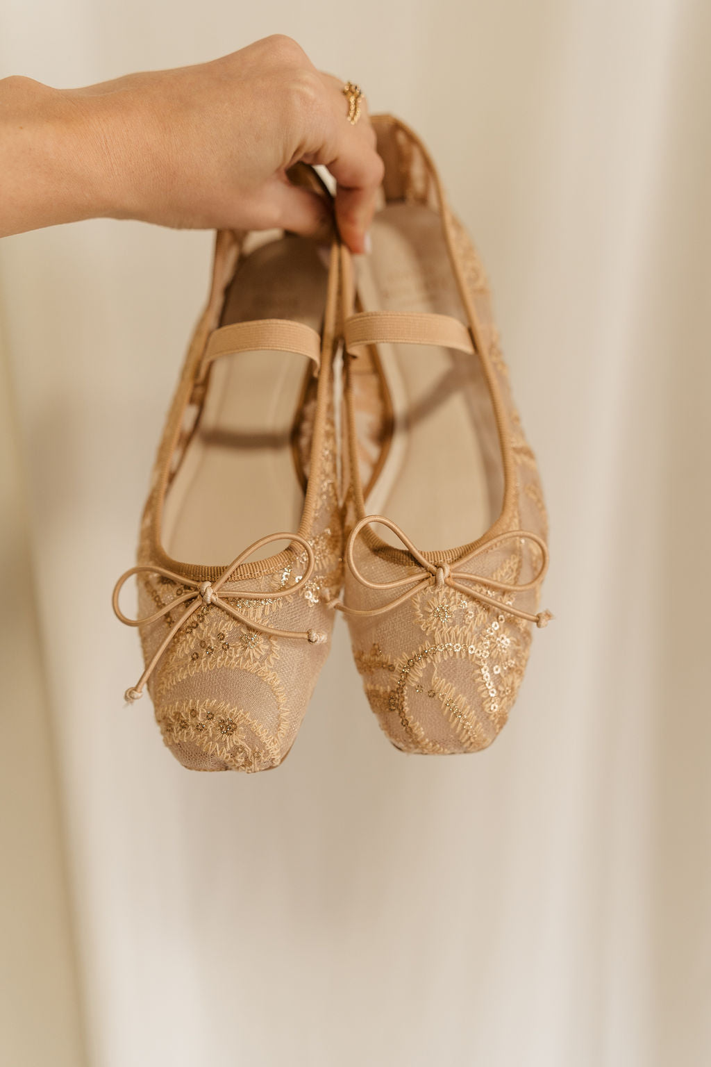 Front view of hand that is holding the Bisbee Ballet Flat in Gold that have beige mesh, ivory embroidery, gold sequins, a bow on front, and an elastic strap across the foot.