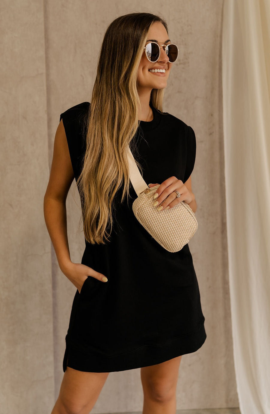 Front view of female model wearing the Rayven Black Sleeveless Mini Dress which features black cotton fabric, mini length, ribbed hem, small side slits, two side slit pockets, a round neckline, and sleeveless.