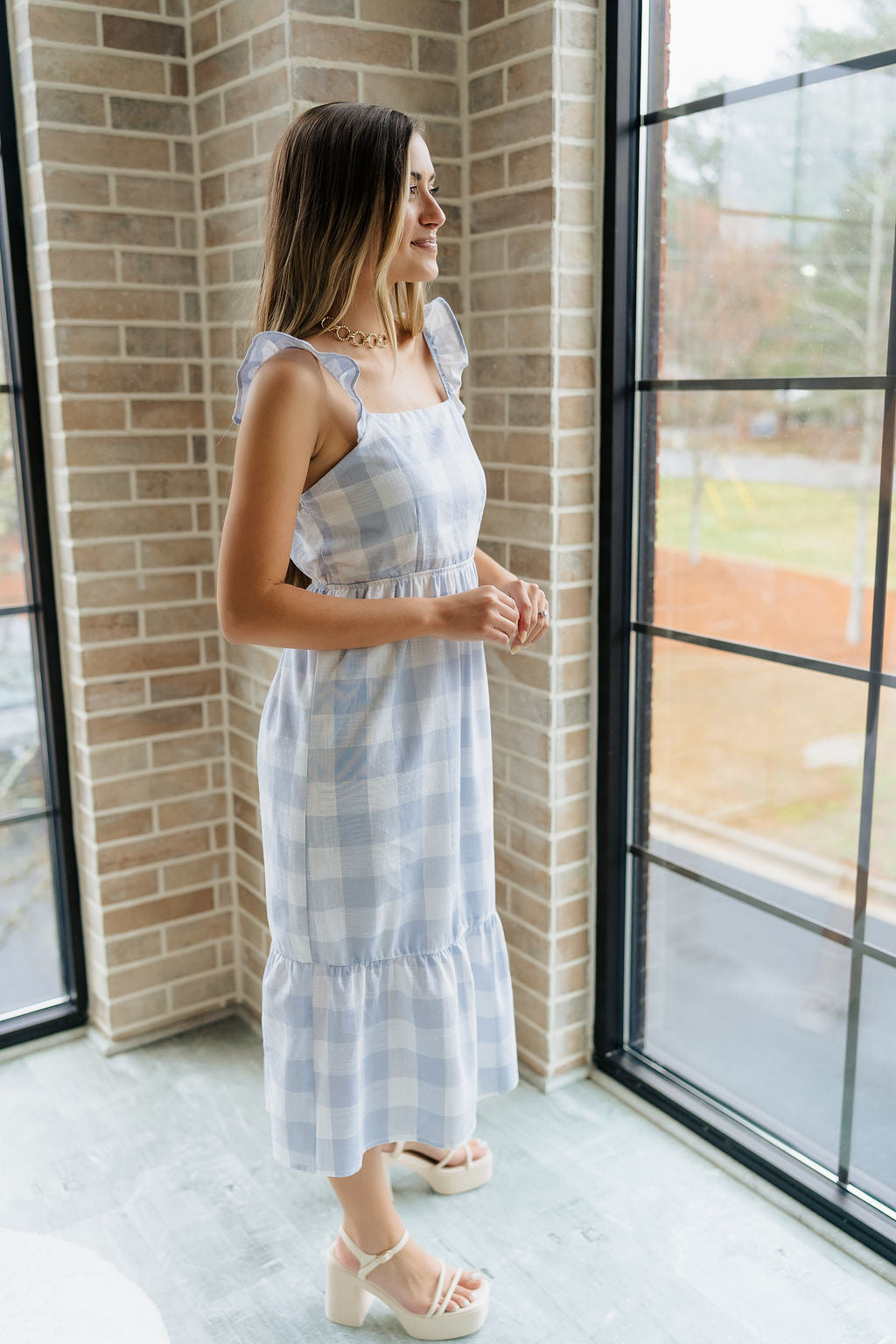 Full body side view of female model wearing the Bonnie Light Blue and White Plaid Midi Dress which features Light Blue and White Plaid Design Midi Length, White Lining, Tiered Ruffle Hem, Square Neckline with Ruffle Straps and Sleeveless.