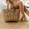 Front view of female model wearing the Beach Please Pearl Woven Tote which features tan rattan woven fabric, "Beach Please" is delicately crafted in pearls on the front and two straps.