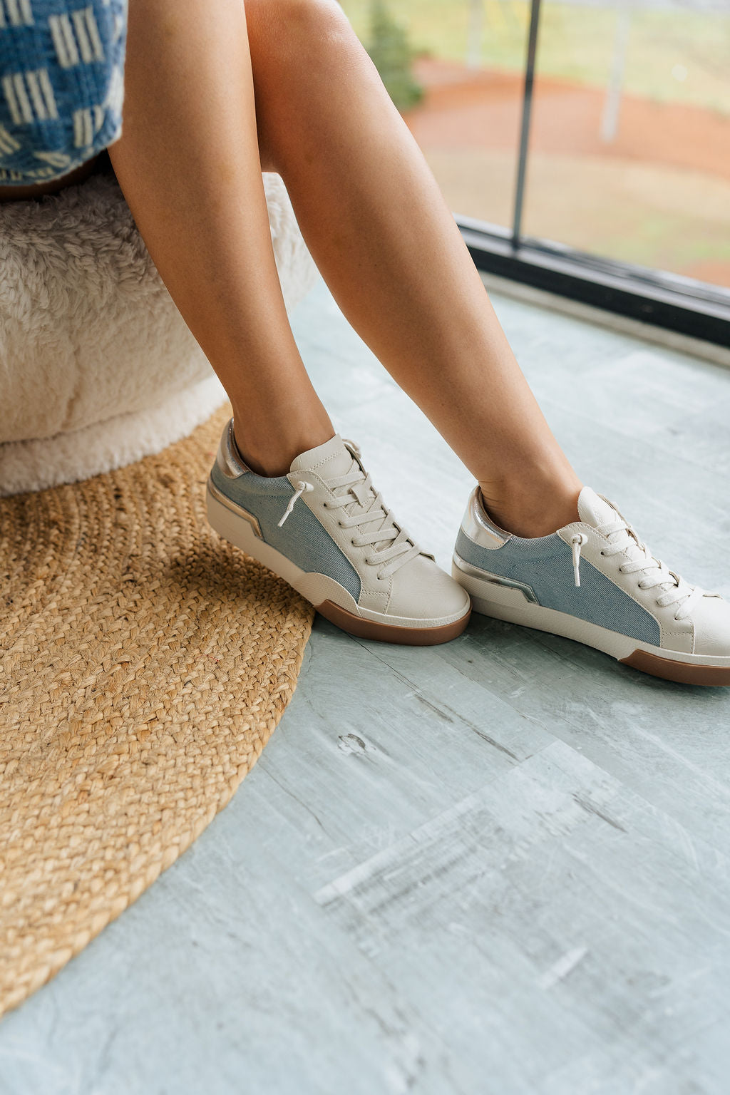 Side view of female model wearing the Helix Sneaker in Denim which features Light Wash Denim Fabric & White Faux Leather Fabric,  Silver Metallic and Light Brown Details, Lace-Up Details and Round Toe.
