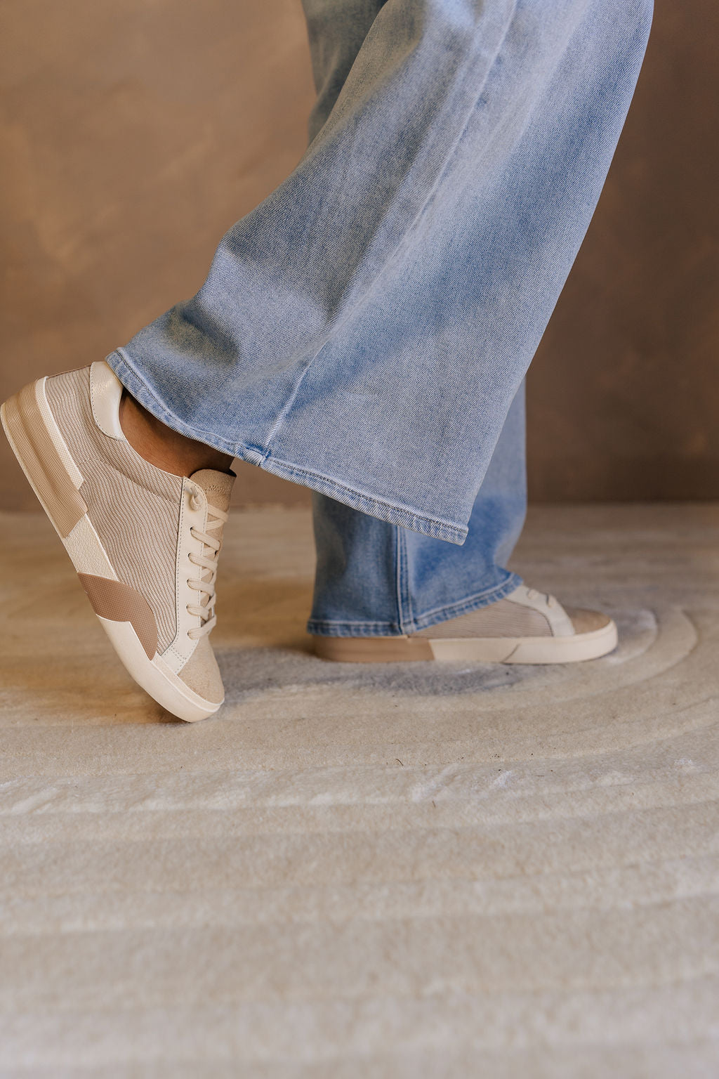 Side view of model wearing the Zina Sneaker in White & Dune Embossed Leather which features white and neutral leather upper fabric, nude suede fabric, lace up closure and round toe.