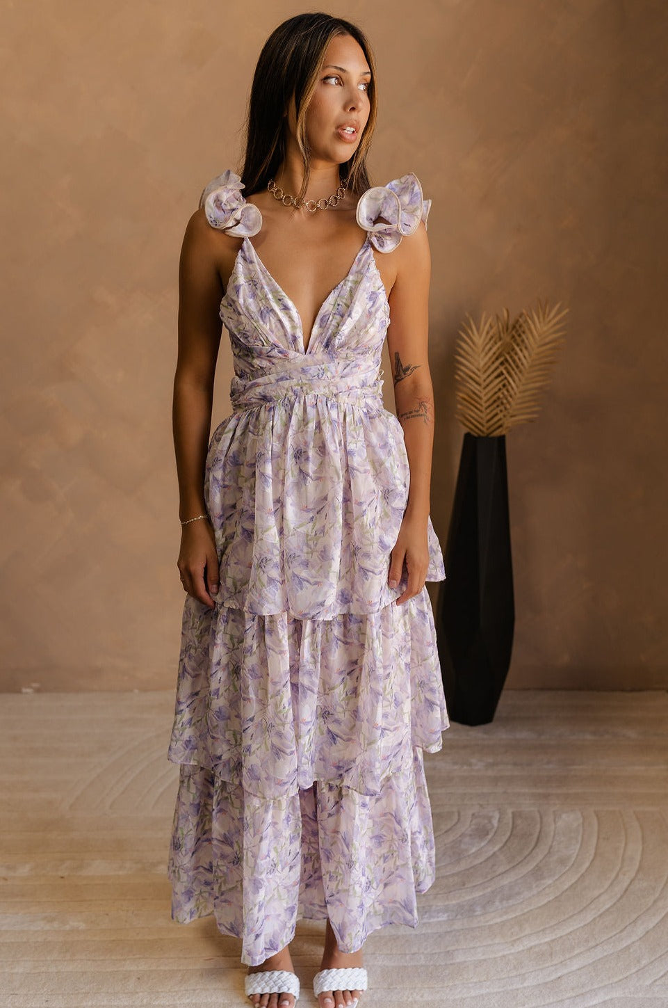 full body view of model wearing The Harlow Lavender Floral Ruffle Midi Dress features sheer fabric with purple, green, orange floral print, shimmer thread details, beige lining, a tiered body with a maxi-length hem, ruffled straps, a plunge neckline, a lace-up back, and a back zipper and hook closure.
