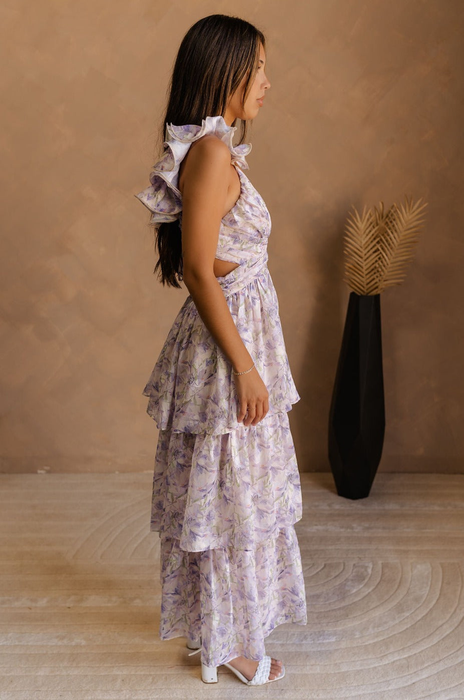 full body side view of model wearing The Harlow Lavender Floral Ruffle Midi Dress features sheer fabric with purple, green, orange floral print, shimmer thread details, beige lining, a tiered body with a maxi-length hem, ruffled straps, a plunge neckline, a lace-up back, and a back zipper and hook closure.