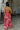 Full body back view of model wearing the Roselyn Pink & Red Floral Maxi Dress that has light pink fabric with dark pink and red floral pattern, a cowl neckline, and spaghetti straps that cross in the back.