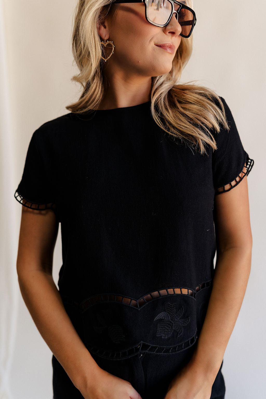 Close up front view of female model wearing the Stella Black Floral Trim Short Sleeve Top which features Black Knit Fabric, Cropped Waist, Scalloped Hem with Open Details, Monochrome Floral Print, Short Sleeves, Round Neckline and Back Button Closure