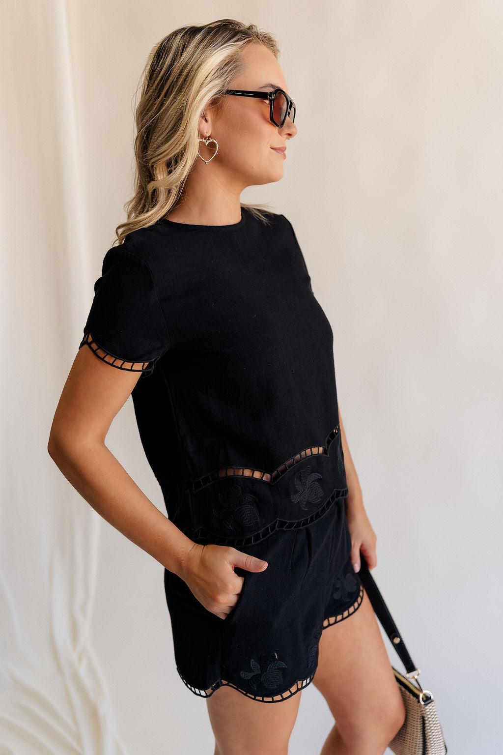 Side view of female model wearing the Stella Black Floral Trim Shorts which features Black Knit Fabric, Scalloped Hem with Open Details, Monochrome Floral Print, Front Zipper with Button Closure, Two Front Pockets and Belt Loops