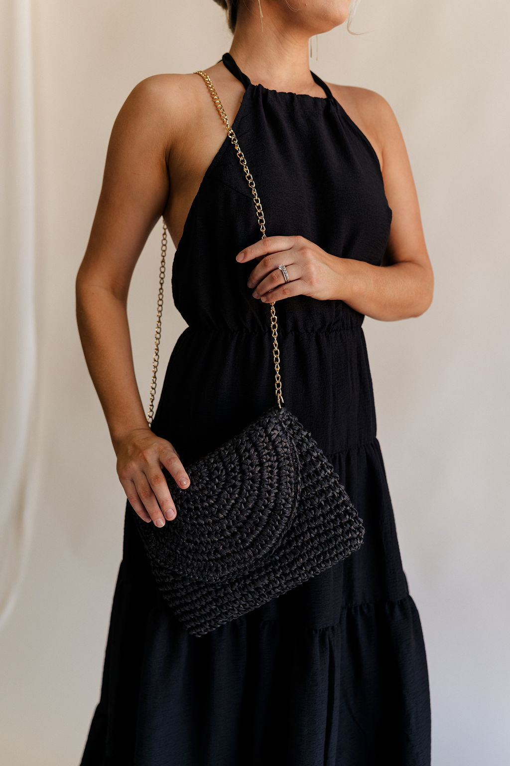 Model is wearing on shoulder the Sofia Black Woven Raffia Purse that has black raffia, a foldover closure with a magnet, and a gold crossbody strap.