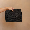 Model's hand is holding the Sofia Black Woven Raffia Purse that has black raffia, a foldover closure with a magnet, and a gold crossbody strap.